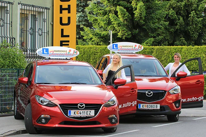 Zwei rote Mazda-Fahrschulautos mit Dachschild vor der Fahrschule Janeschitz, daneben zwei Fahrlehrerinnen